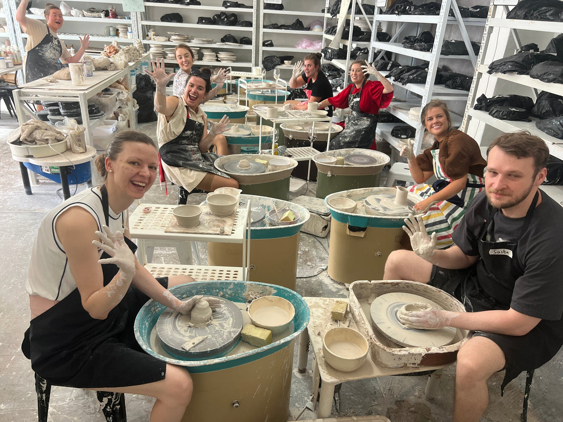 Group of people sitting aorund pottery wheels and making pottery, smiling and waving at the camera